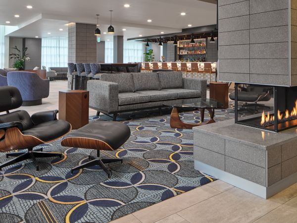 A modern, upscale hotel lounge with a fireplace, Eames lounge chair, gray sofas, patterned rug, and a bar in the background.