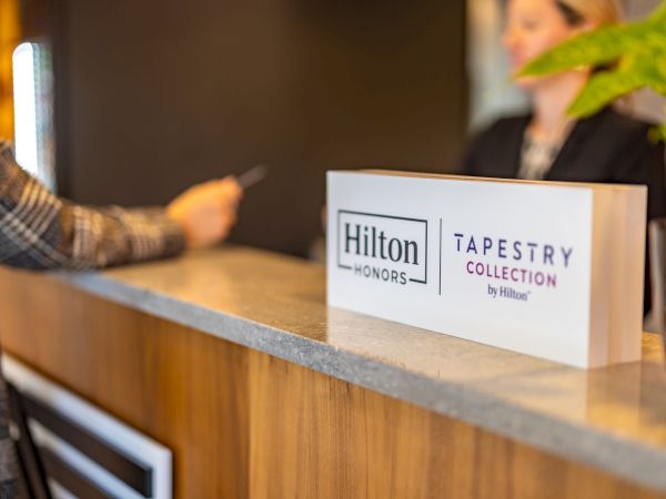 A hotel reception desk with a &ldquo;Hilton Honors&rdquo; sign and a &ldquo;Tapestry Collection by Hilton&rdquo; display, blurred guest in background, welcoming staff.