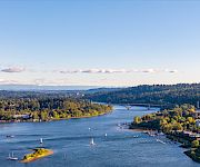 A wide river flows past forested hills, with sailboats, a marina, and a bridge stretching across under a blue sky.
