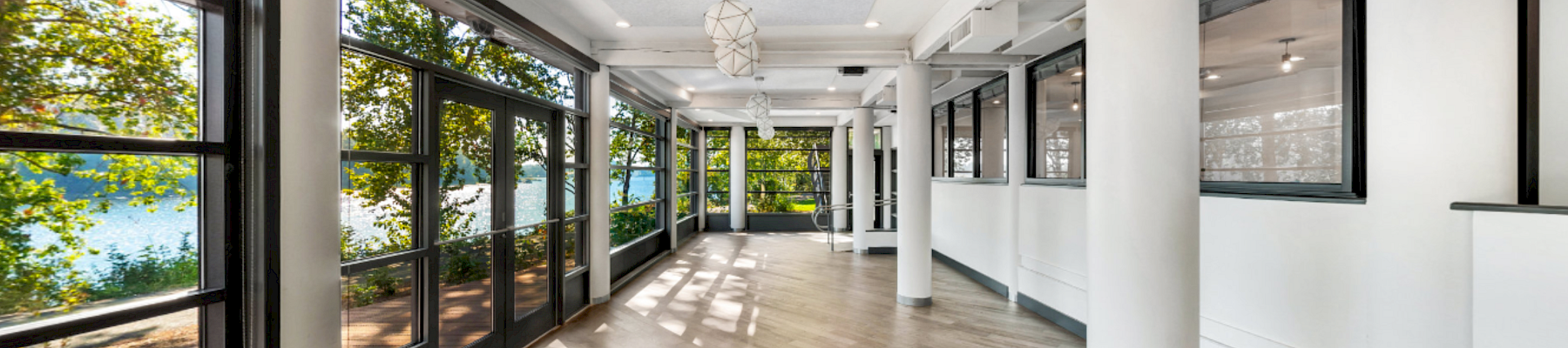 Bright modern hallway with floor-to-ceiling windows, white columns, wood floors, and hanging pendant lights.