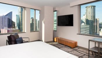 A modern hotel room features large windows with city views, a bed, a TV, and contemporary furniture.