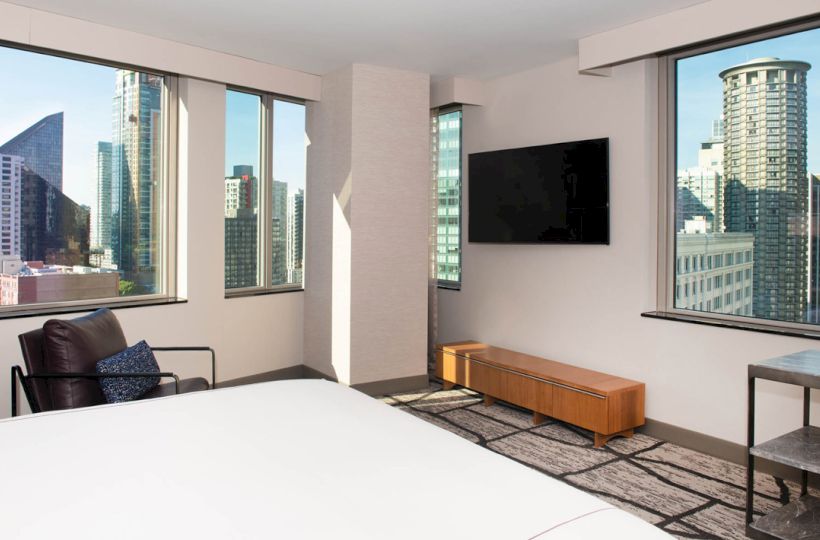A modern hotel room features large windows with city views, a bed, a TV, and contemporary furniture.