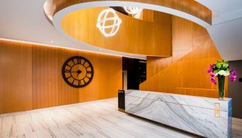 The image shows an elegant lobby with a marble reception desk, a large wall clock, modern lighting, and a wooden spiral staircase.
