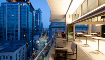 A modern rooftop terrace with seating, overlooking a city street lined with tall buildings at dusk, softly illuminated by ambient lighting.