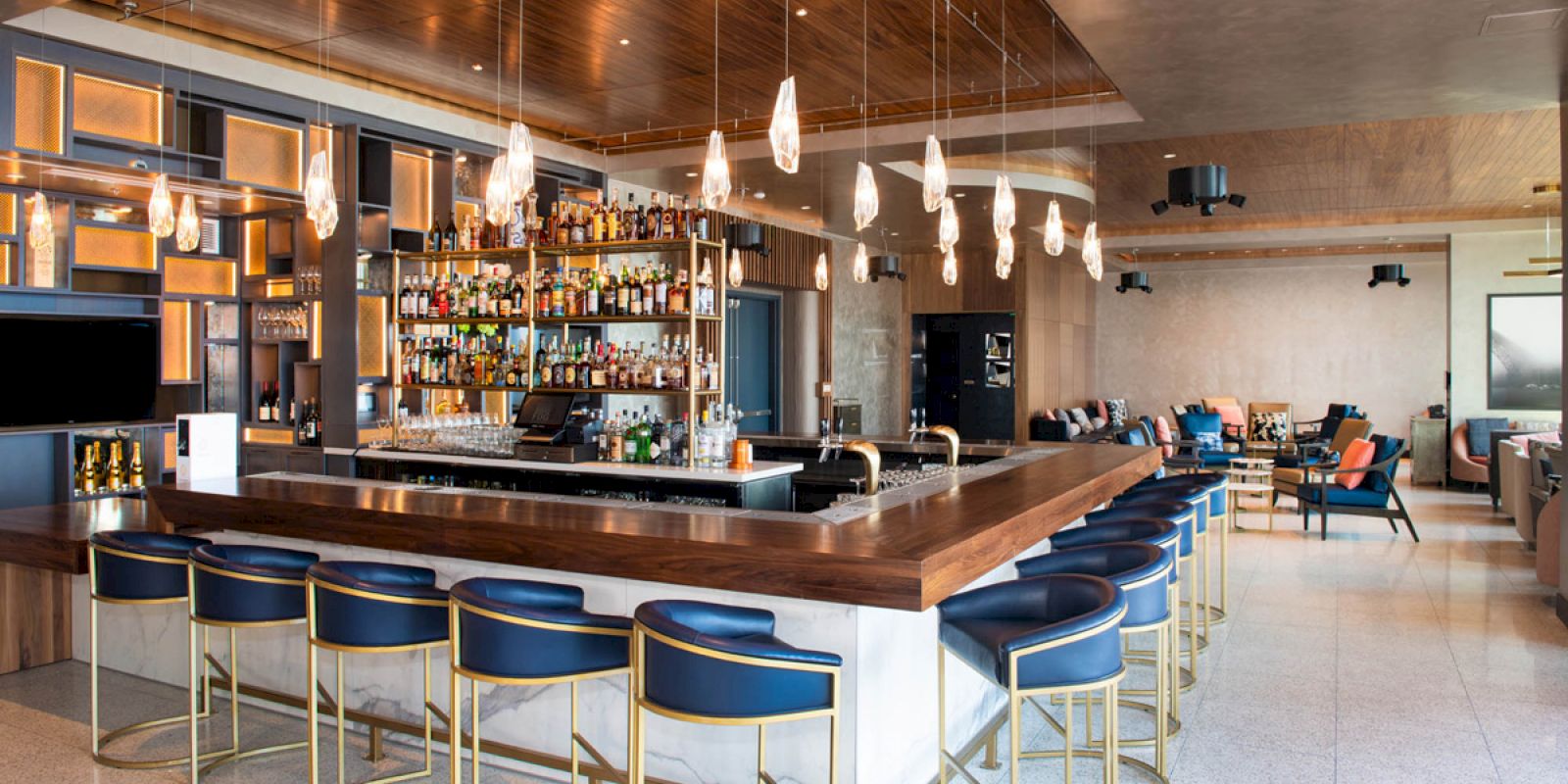 The image shows a modern bar with blue barstools, hanging lights, and a well-stocked liquor shelf, in a stylish lounge area.