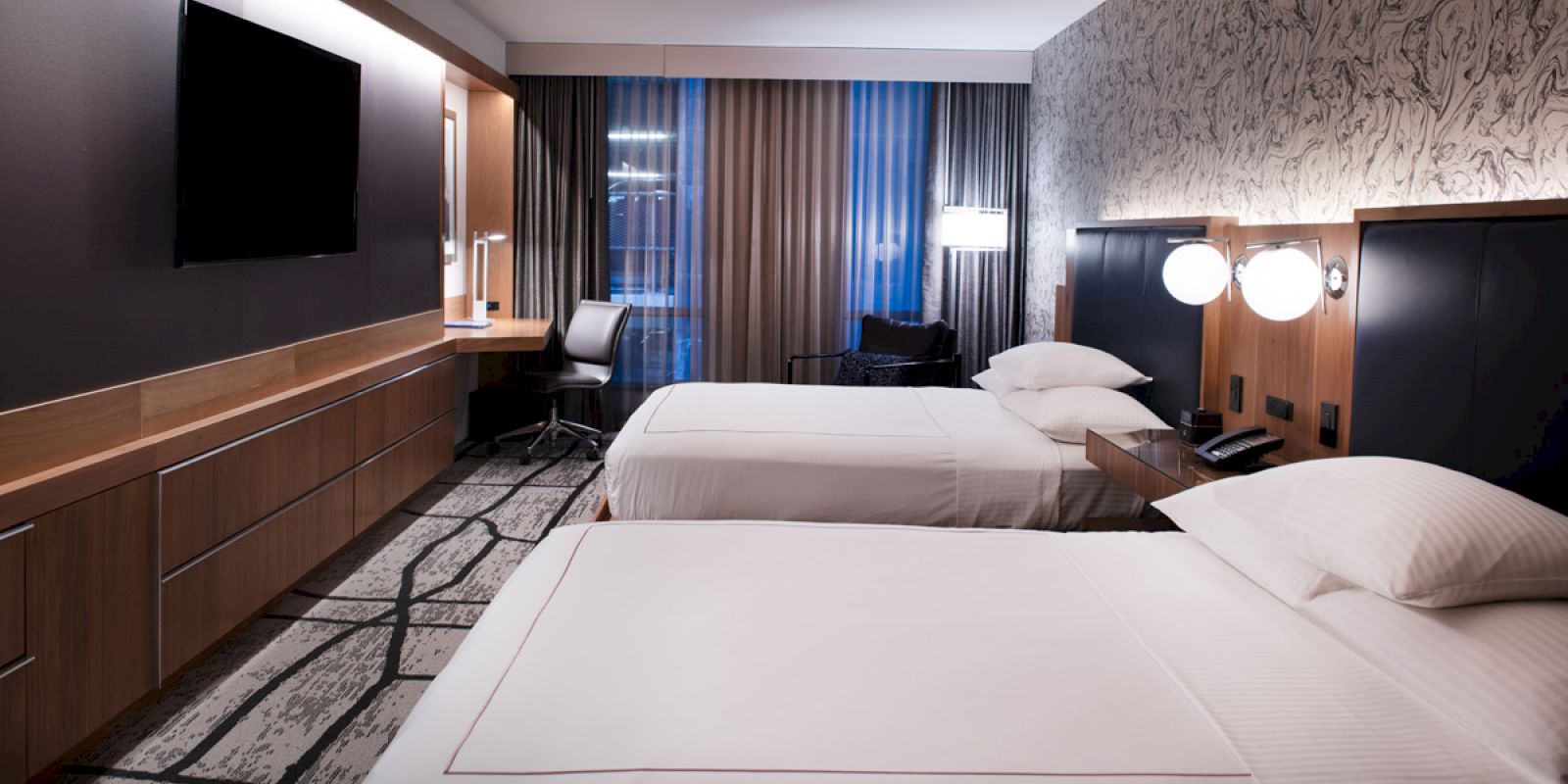 The image shows a modern hotel room with two double beds, a wall-mounted TV, a desk with a chair, and contemporary decor.