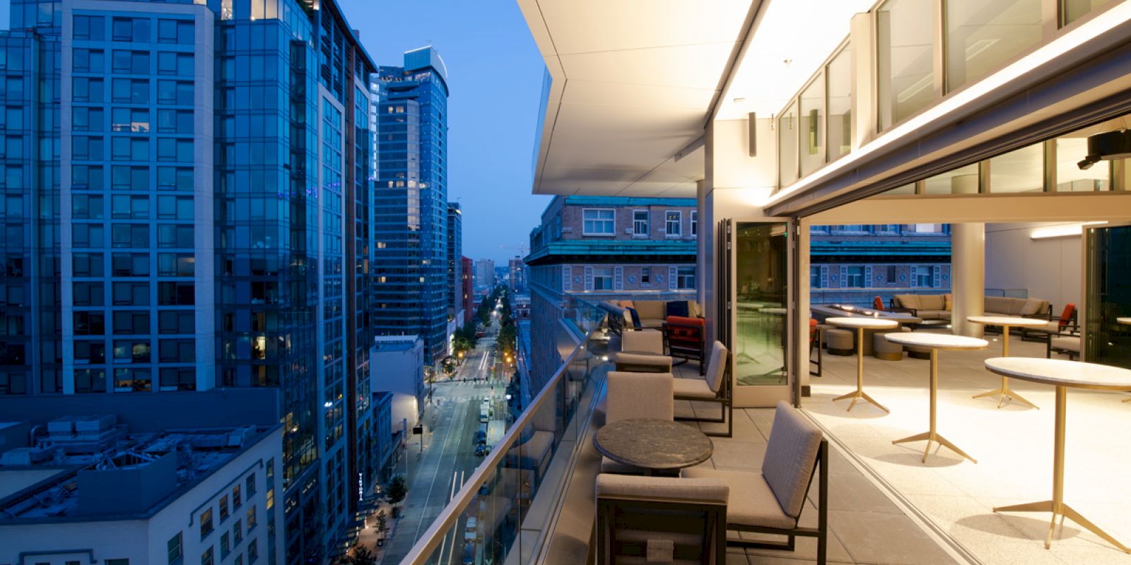 A modern high-rise balcony overlooks a cityscape at dusk, with tables and chairs along the rail and buildings illuminated in the distance.