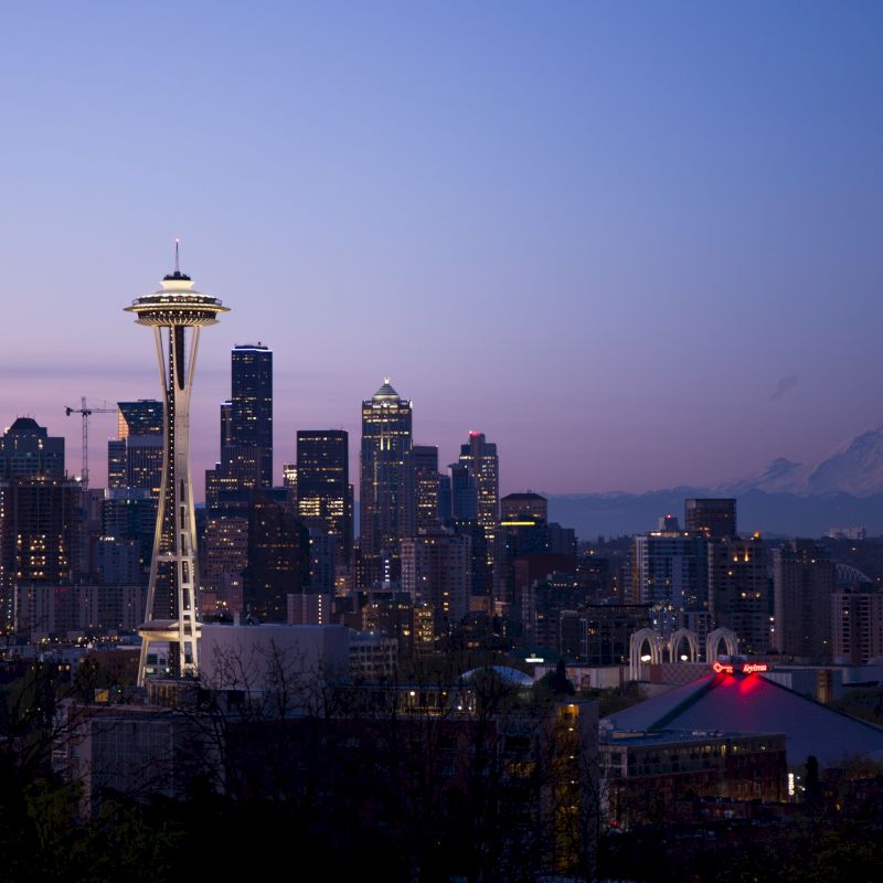 Seattle skyline at dusk with the Space Needle prominently visible and Mount Rainier in the background, under a clear sky.
