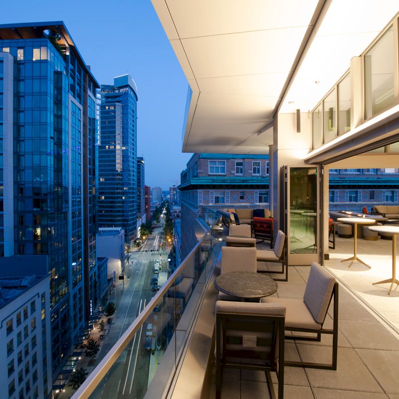 The image shows a modern cityscape view from a balcony, with tables and chairs, overlooking tall buildings at dusk.