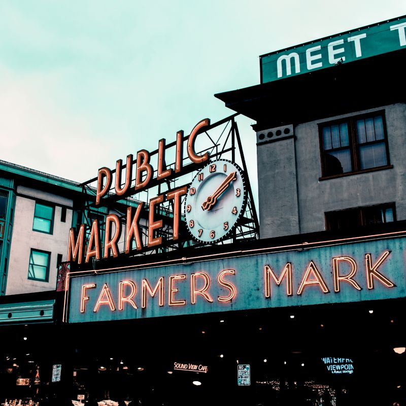 The image shows a vibrant neon sign for a "Public Market" and "Farmers Market" with buildings in the background under a cloudy sky.