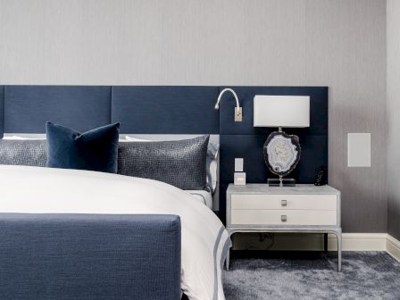 A modern bedroom with a navy headboard, white bed, and side table with a lamp and decor. Plush carpet adds comfort to the space.