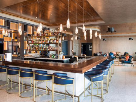 A modern bar with blue stools, hanging lights, and a stocked drink shelf. There's seating in the background, creating a cozy atmosphere.