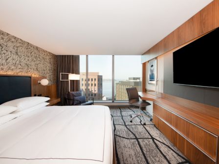 A modern hotel room features a large bed, a desk with a chair, and a big TV, with a city view through the window.