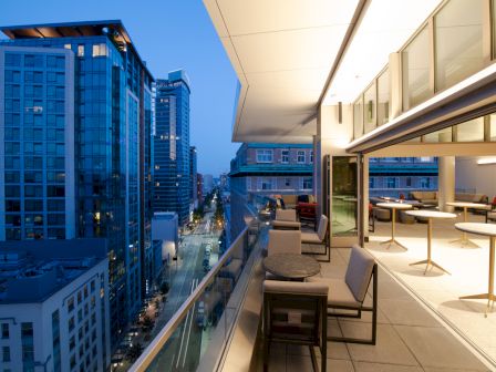 A cityscape at dusk featuring a modern balcony with seating and large buildings lining both sides of a street.