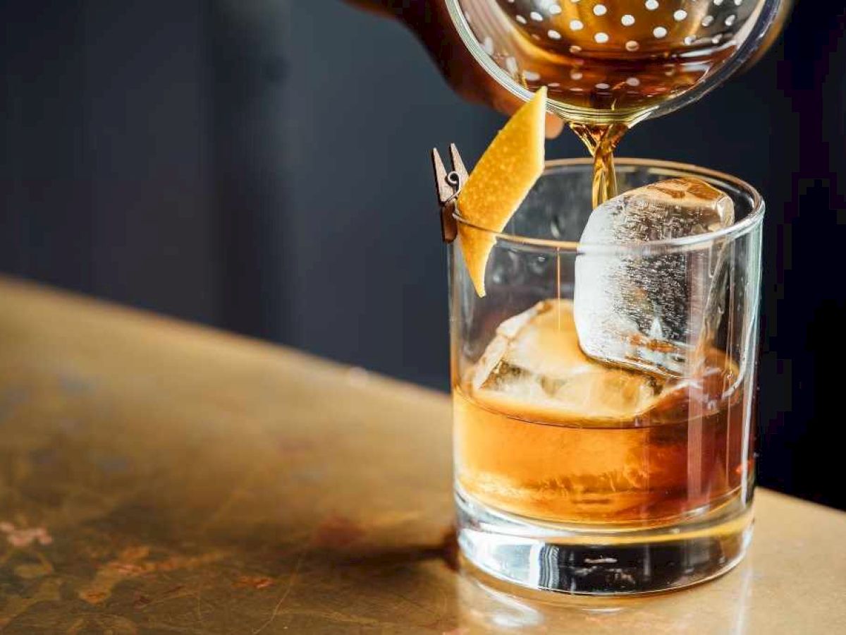 A classic cocktail being poured into a glass with ice and a lemon twist garnish on a bar counter.