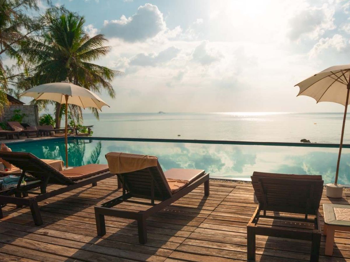 A serene beach scene with lounge chairs, umbrellas, and a pool overlooking the ocean, surrounded by palm trees under a partly cloudy sky.