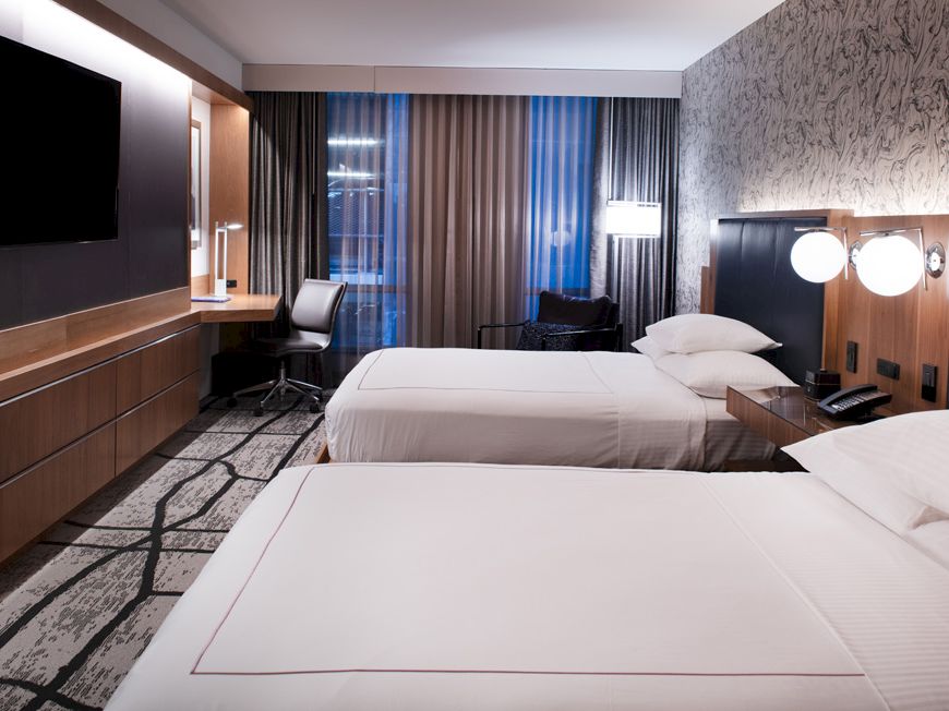 The image shows a modern hotel room with two beds, a wall-mounted TV, a desk with a chair, stylish lighting, and patterned carpet and wallpaper.