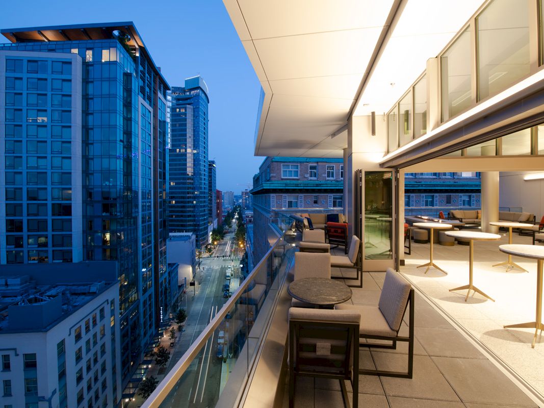 A modern rooftop patio with seating overlooks a city street lined with tall buildings at dusk.