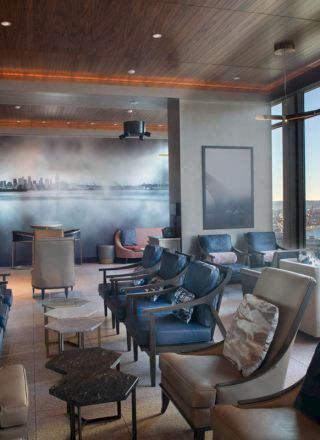 A modern lounge with large windows offers a scenic view, comfortable seating, and a city mural on the wall, with a waterfront outside.