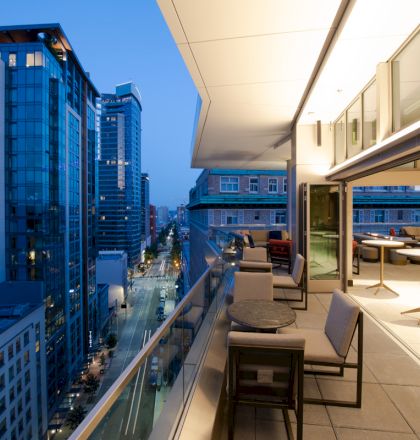 A modern rooftop patio with tables and chairs overlooking a cityscape with tall buildings at dusk.