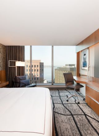 The image shows a modern hotel room with a bed, desk, chair, large TV, and window view of buildings and water in the background.