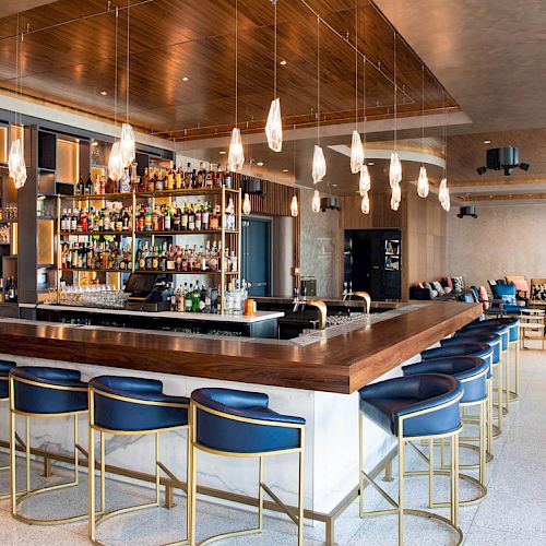 The image shows a modern bar with a wooden counter, blue bar stools, hanging lights, a stocked liquor shelf, and cozy seating in the background.