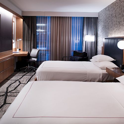 A modern hotel room with two beds, a desk, a TV, and stylish decor, featuring patterned wallpaper and ample lighting.