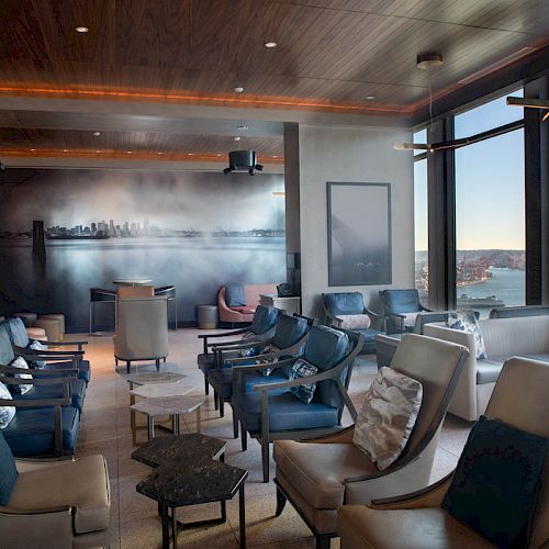 A modern lounge with plush seating, a wall mural, large windows offering a scenic view of the ocean, and a Ferris wheel outside.