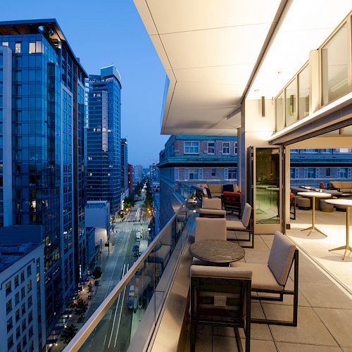 A modern rooftop terrace with tables and chairs, overlooking a cityscape of tall buildings during twilight hours.