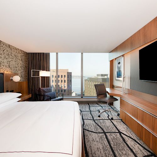 A modern hotel room with a large bed, desk, chair, large TV, and a window view of city buildings and water are in the image.