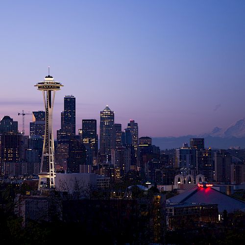 A city skyline at dusk featuring a prominent tower, surrounded by buildings, with a mountain in the background under a clear sky.