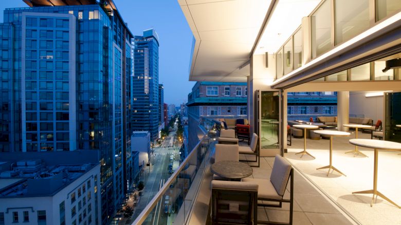 A modern cityscape view from a balcony at dusk, featuring tall buildings, a glass railing, tables, and chairs in an urban setting.