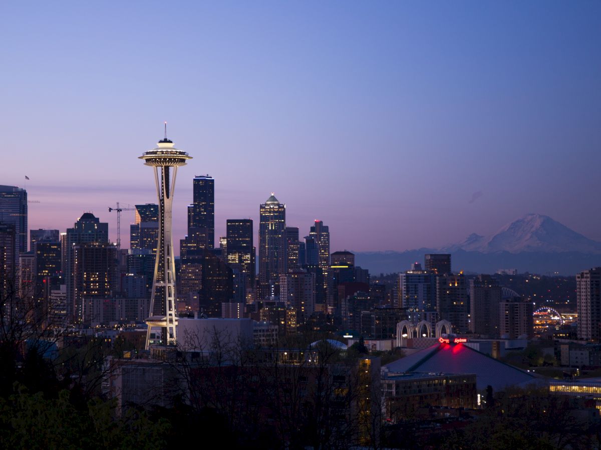 Seattle skyline at dusk, featuring the Space Needle and Mount Rainier in the background, under a clear evening sky.