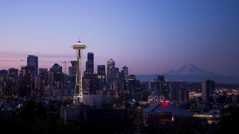 Seattle skyline at dusk, featuring the Space Needle and Mount Rainier in the background, under a clear evening sky.