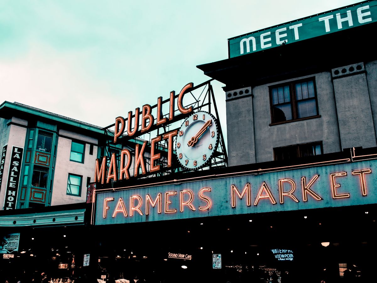 The image shows a public and farmers market sign with a clock, set against a backdrop of buildings, suggesting an urban setting.