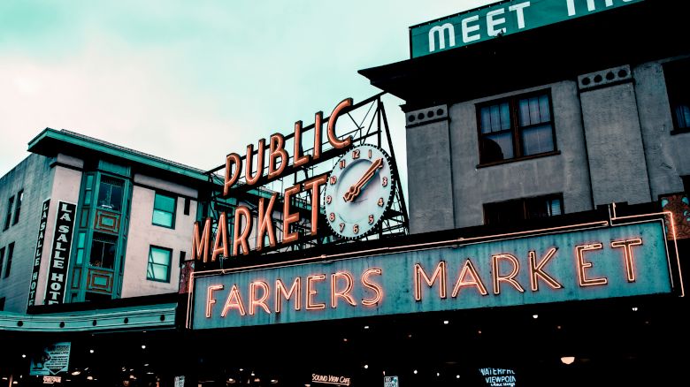 The image shows a public and farmers market sign with a clock, set against a backdrop of buildings, suggesting an urban setting.