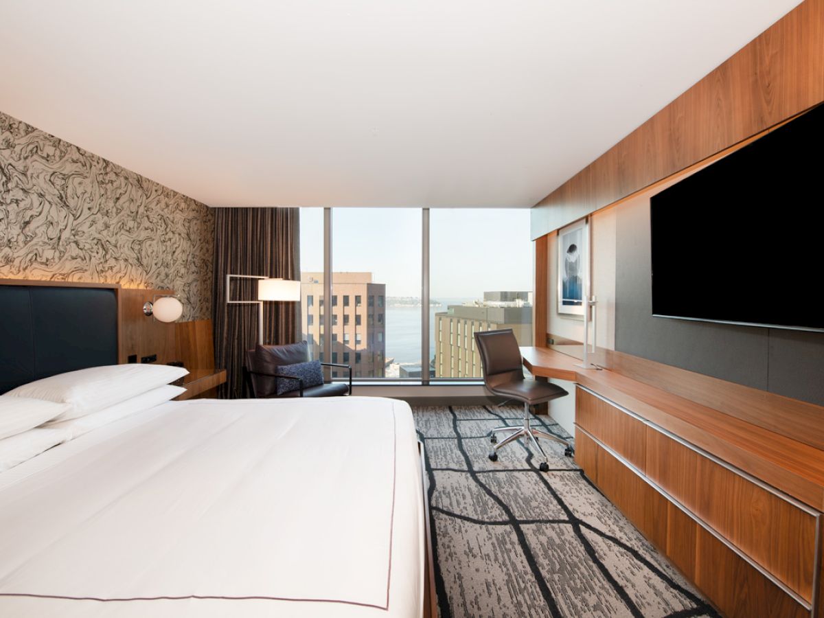 The image shows a modern hotel room with a bed, desk, TV, and a view of city buildings through large windows.