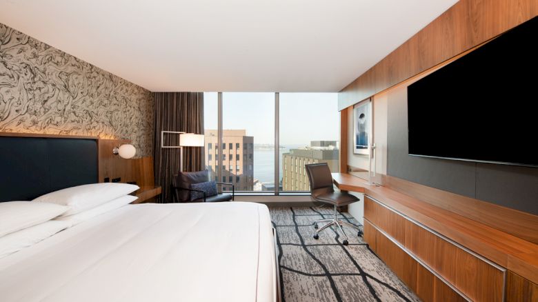 The image shows a modern hotel room with a bed, desk, TV, and a view of city buildings through large windows.