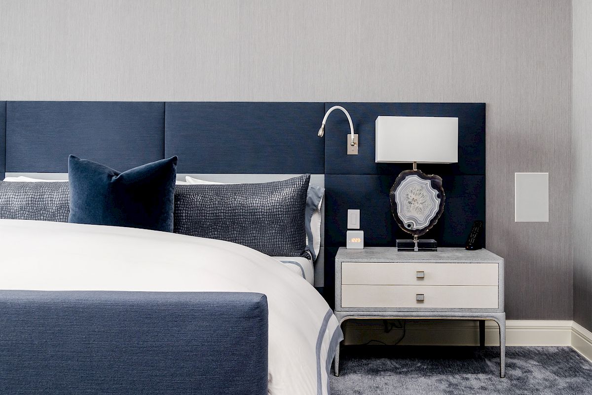 A modern bedroom with a blue-themed design, featuring a bed, pillows, a bedside table with a lamp, and minimalistic decor.
