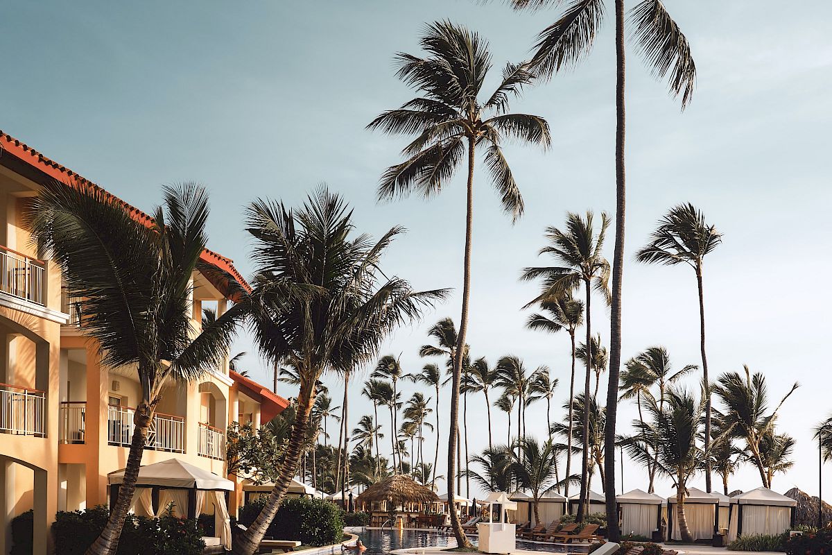 A tropical resort scene features a pool, palm trees, and sun loungers under a blue sky. Relaxing and inviting atmosphere.
