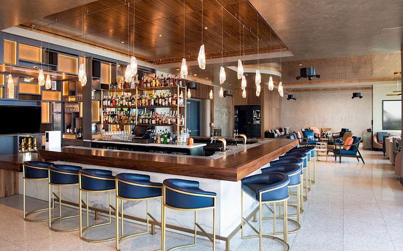 A modern bar with a central island counter, stylish blue stools, pendant lights, a well-stocked back bar, and cozy seating area.
