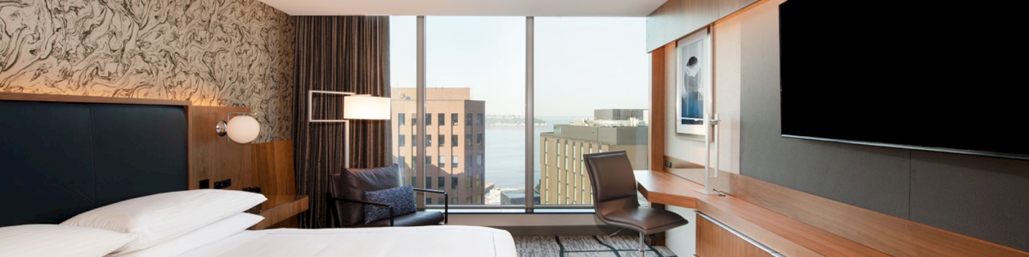 A modern hotel room features a large bed, wall-mounted TV, desk, and a window with a city view, decorated with wood and patterned walls.