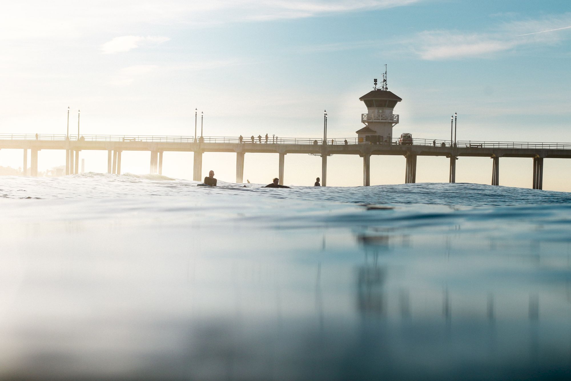 The image shows a pier extending over the ocean with people walking on it, under a clear sky, viewed from the water.